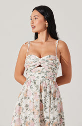 A woman is wearing a sleeveless dress with thin shoulder straps tied into bows. The dress features a fitted bodice with a sweetheart neckline and a center knot detail. The skirt part of the dress is flowy and has a floral pattern with soft pastel colors, including pink, green, and peach, on a light background.